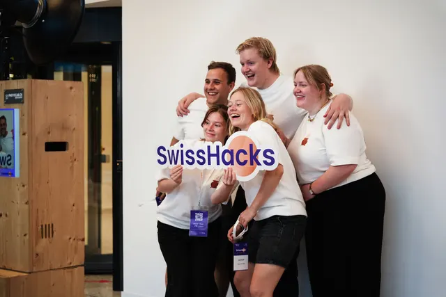 Five participants laughing at the photo booth while holding a cut-out SwissHacks sign.
