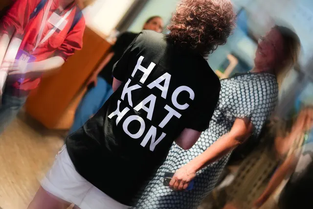 A participant in a black Hackathon shirt seen from behind in a busy room.