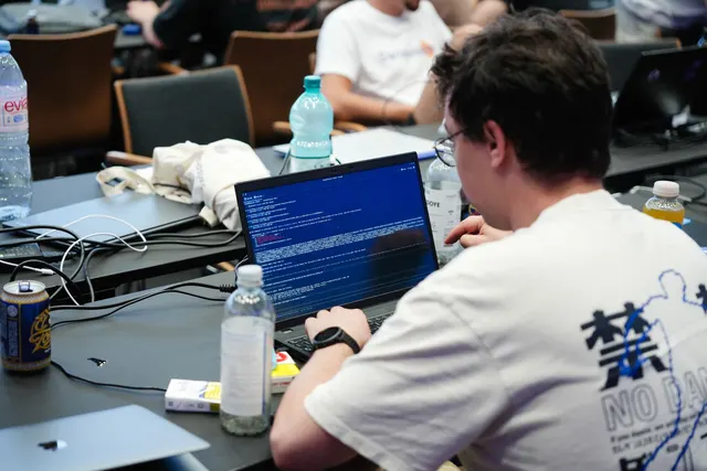 A participant coding on a laptop full of terminal windows, surrounded by drinks and cables.