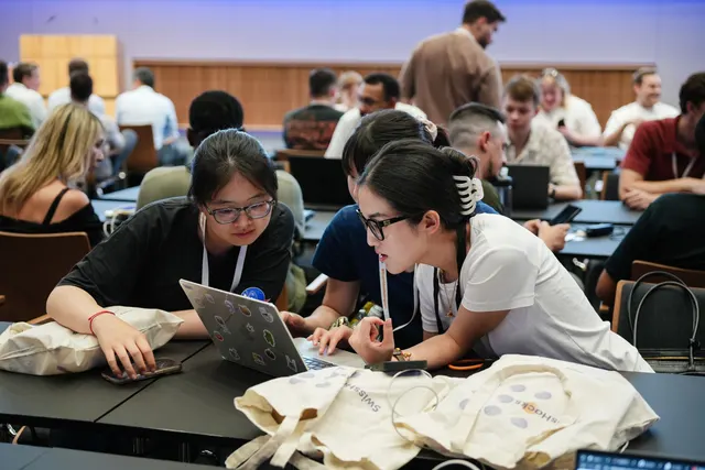 Three participants leaning over one laptop together, SwissHacks tote bags on the table.